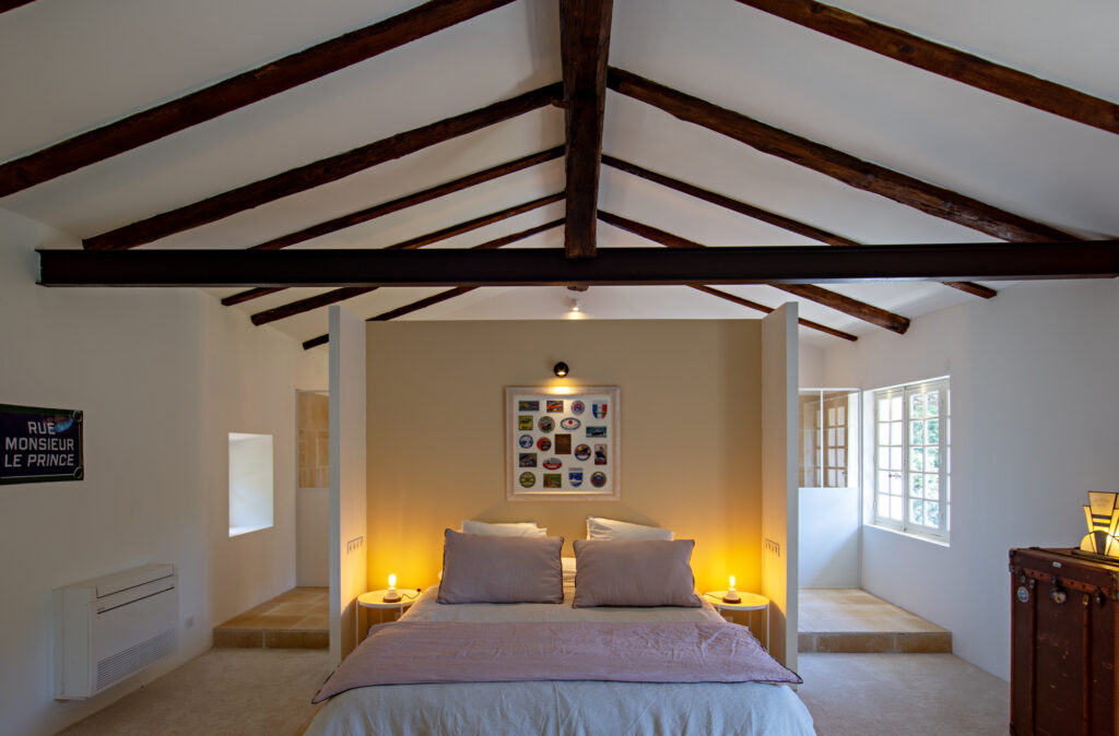Chambre du Mas La Solarie avec lit double, poutres en bois apparentes, tables de chevet &eacute;clair&eacute;es et salle de bain attenante.