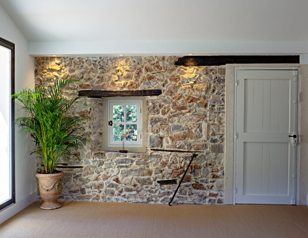 Mur en pierres apparentes avec poutres en bois, petite fen&ecirc;tre et plante d&eacute;corative dans une salle du Mas La Solarie.