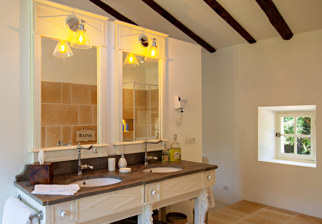Salle de bain du Mas La Solarie avec double vasque, grand miroir, poutres en bois au plafond et petite fen&ecirc;tre donnant sur la nature.