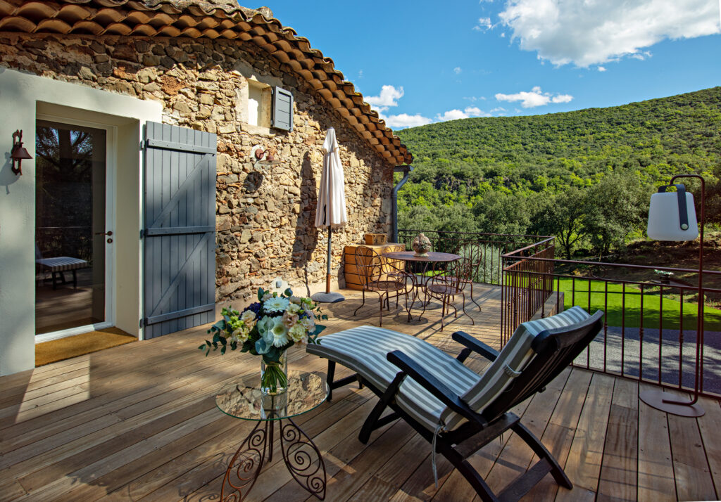 Terrasse du Mas La Solarie avec mur en pierres apparentes, table de jardin, transat et vue panoramique sur la nature et les collines.